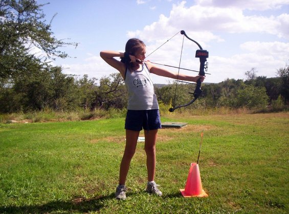 Girl aims bow and arrow at target in grassy field