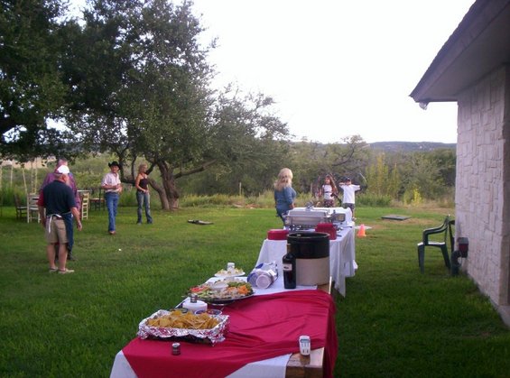 Outdoor buffet table set up on a lawn with people standing around, trees in the background