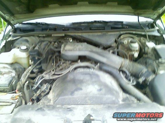 Engine bay with various hoses and components, dusty and worn. Water reservoir visible on the left