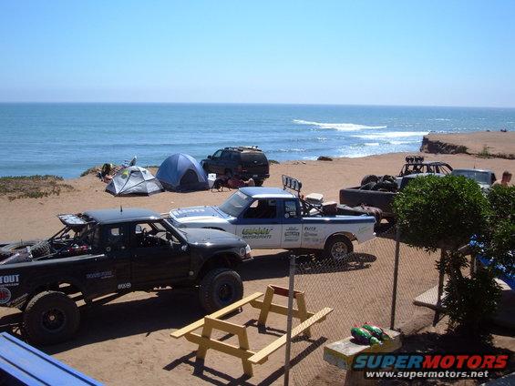 Several off-road trucks, tents, and picnic tables are set up on a sandy beach with the ocean in the background