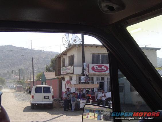 White van parked on a dirt road, rear view. Buildings and people in the background