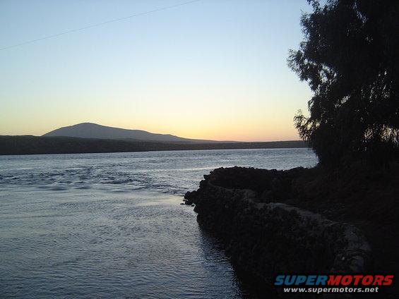 River flows past a rocky shore with a tree silhouette against a sunset sky and distant mountains
