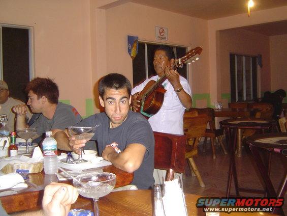 guy holding a margarita looks at the camera while a musician plays guitar behind him