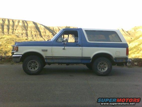 Two-tone blue and white Ford Bronco parked on a road with a rocky hillside in the background