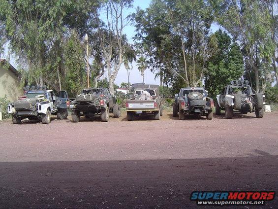 Five modified off-road trucks parked in a dirt lot with trees in the background