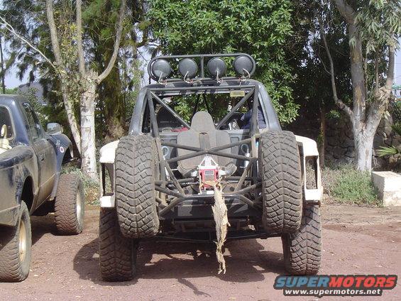Off-road buggy rear view, white body, roll cage, four lights on roof, two spare tires, one mounted vertically