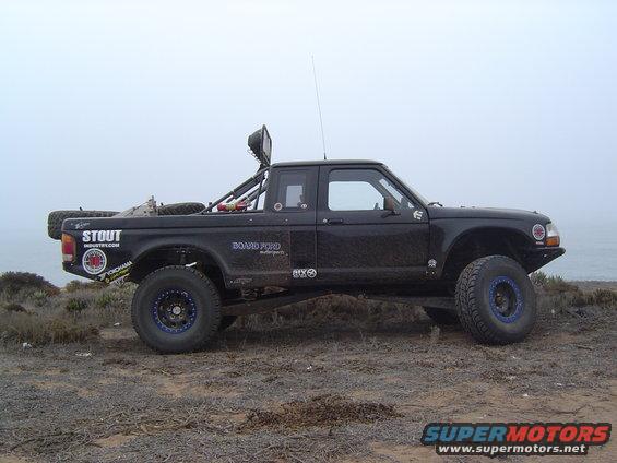 Black off-road pickup truck with large tires and a roll cage, parked on a dirt hill near the ocean