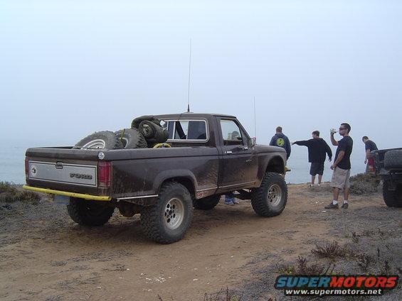 Brown Ford Ranger pickup truck parked on a dirt cliff overlooking the ocean, spare tires in the bed