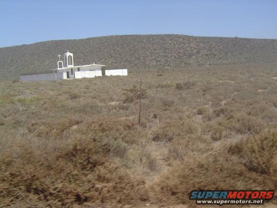 White church building with a bell tower sits on a hill in a dry, scrubby landscape under a clear blue sky