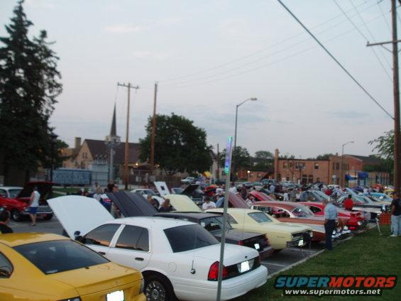 Cars lined up at a car show, some with hoods open. A church steeple is visible in the background