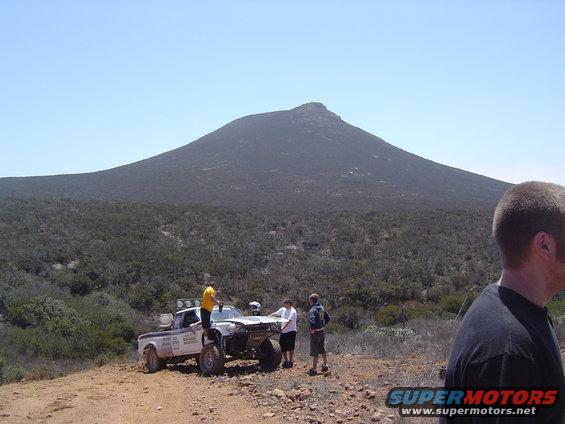 White off-road pickup truck with large tires, modified front end, and people gathered around, parked on a dirt track with a mountain in the background