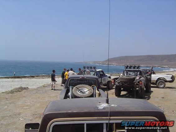 Several off-road trucks are parked on a dirt lot near the ocean, with people standing around them