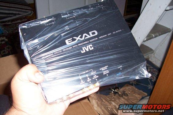 Hand holding a JVC EXAD KD-AV7010 multimedia DVD/CD receiver with removable monitor, still in plastic wrap