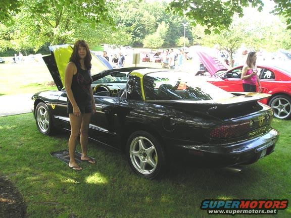 Woman standing next to a black Pontiac Firebird Trans Am with its hood up. A red car is parked nearby