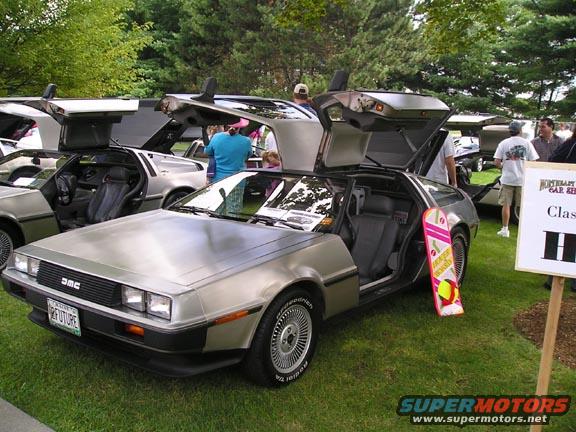 Several DeLorean DMC-12 cars with gullwing doors open, parked on grass. One has a "Back to the Future" themed hoverboard leaning against it. A sign reads "MONTGOMERY CAR SHOW Class H"