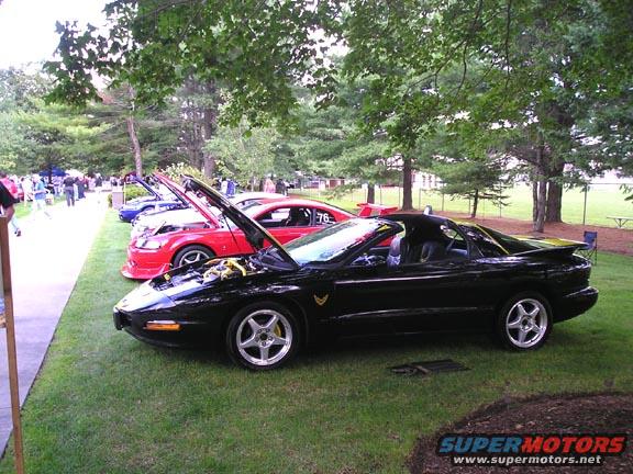Black Pontiac Firebird parked on grass at car show, hood open. Red car next to it also has hood open