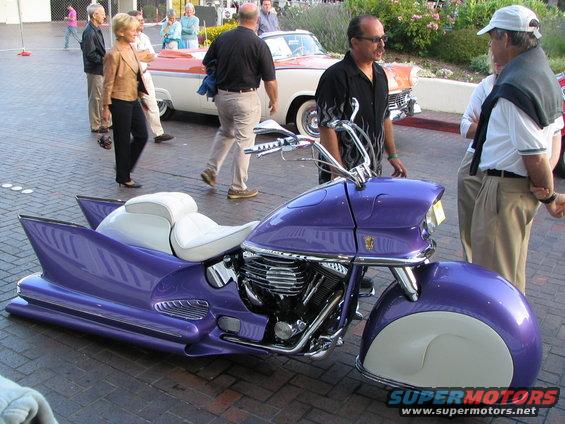 Custom purple motorcycle with white seats and large fins, parked on brick pavement. Two classic cars and people are in the background