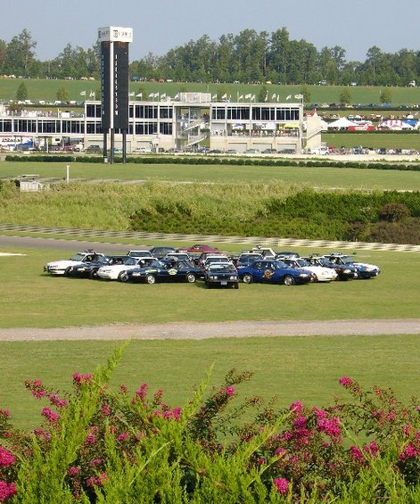 Cars parked on grass at a racetrack with grandstands in the background