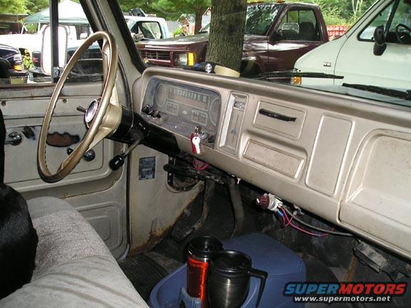 Old truck interior showing dashboard, steering wheel, and gauges. Other vehicles visible outside