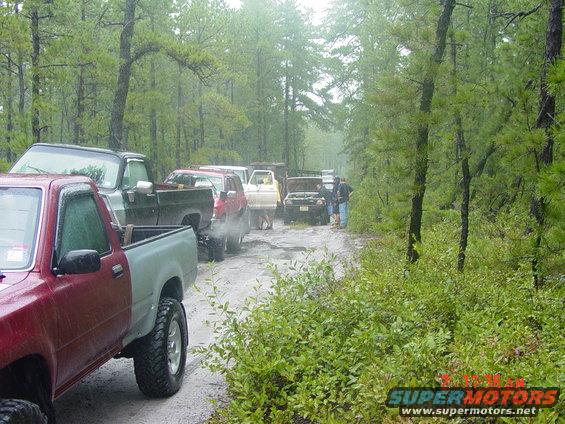 Line of off-road vehicles on a muddy dirt road in a forest, some with hoods open