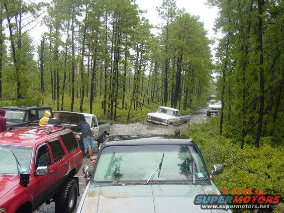 Several off-road vehicles navigate a muddy path through a forest; one white pickup truck is partially submerged in water