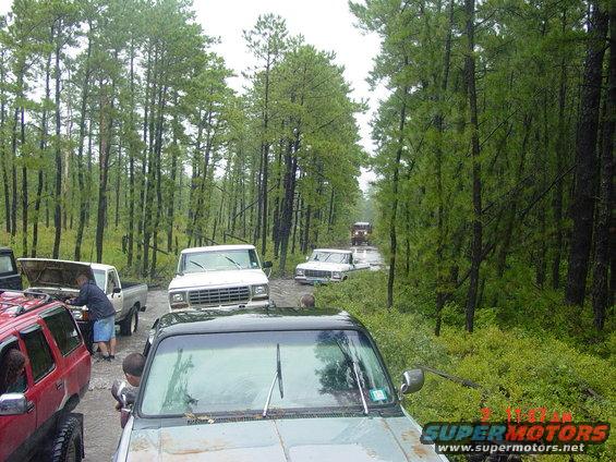 Several trucks are stuck in mud on a wooded road. One truck has its hood open