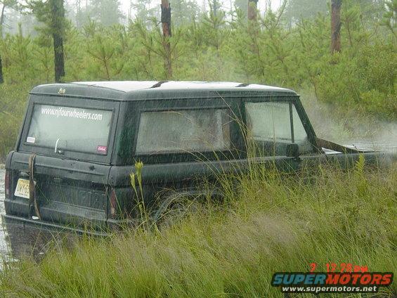 Dark green SUV partially submerged in water, surrounded by tall grass. Website www.njfourwheelers.com visible on rear window