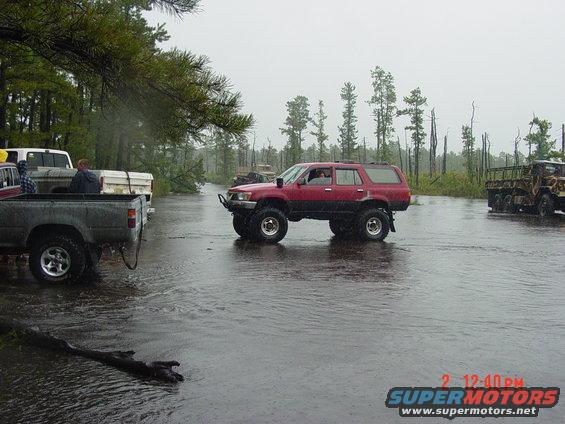 Red SUV with lifted suspension driving through water, other trucks visible