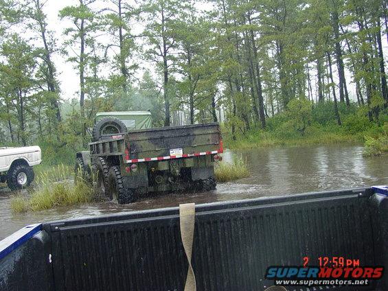 Military truck with camouflage paint driving through a flooded area with trees in the background. Another white truck is visible to the left