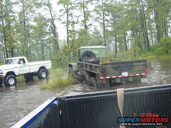Rear view of a camouflaged military truck driving through water, with a white pickup truck behind it