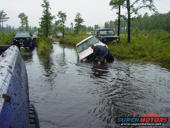 White van stuck in water, person pushing. Other trucks visible