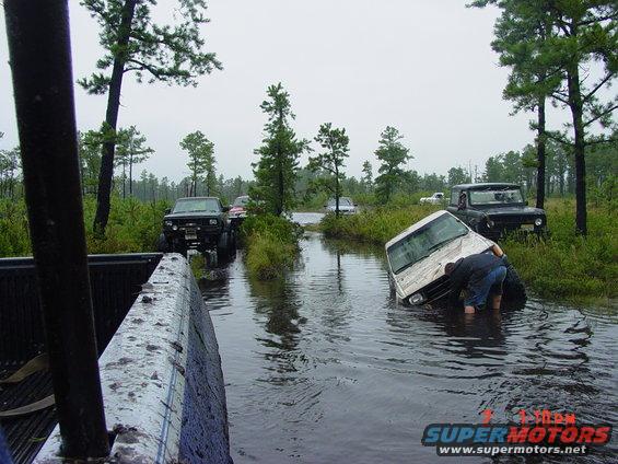 Several off-road vehicles are stuck in a flooded area with trees and brush around. One person is checking a white vehicle that is partially submerged