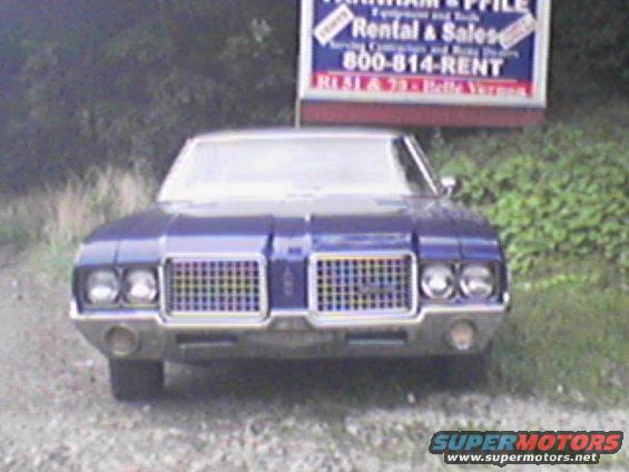 Front view of a blue 1973 Oldsmobile Cutlass with a large chrome grille and quad headlights. Sign in background reads 'Rental & Sales'