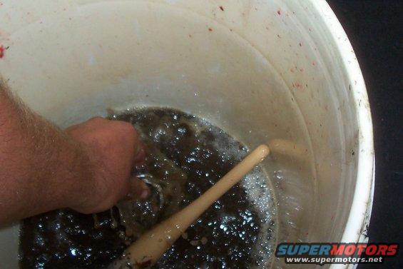 Hand stirring murky liquid in a white bucket with a wooden stick