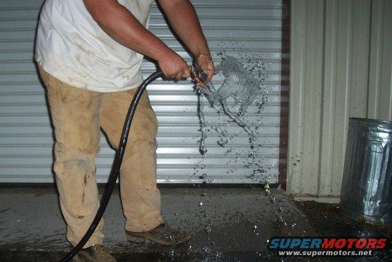 Person in tan pants and white shirt spraying water from a hose in front of a metal garage door. A metal trash can is to the right