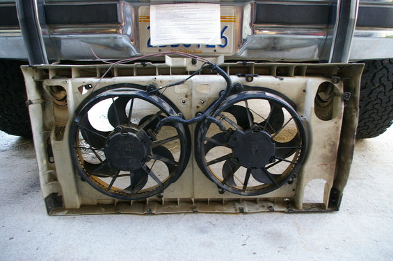Dual electric fans mounted in a plastic shroud, with a vehicle bumper and license plate visible above