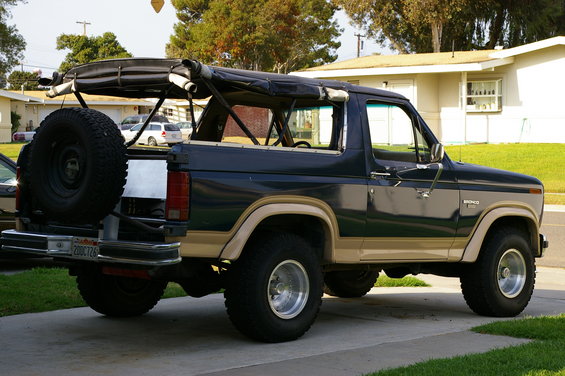 Side view of a dark blue and tan Ford Bronco with a soft top frame visible. Spare tire mounted on the rear. License plate reads 200CT26