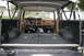 1986 Ford Bronco - The interior of a Ford Bronco shows a brown dashboard and a black spray-lined cargo area, with the rear tailgate open.