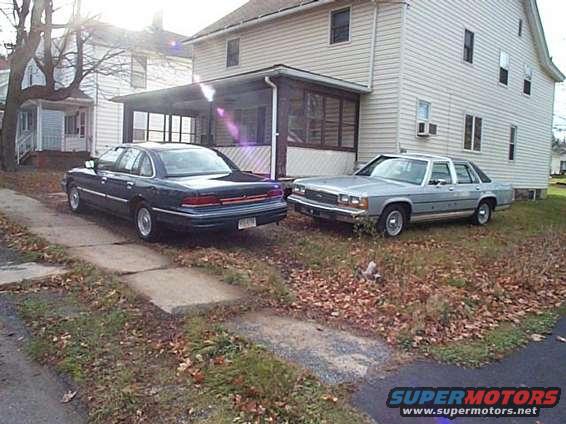 Dark blue sedan and a light blue sedan parked in front of a house. The blue sedan is a 1991 Ford Crown Victoria