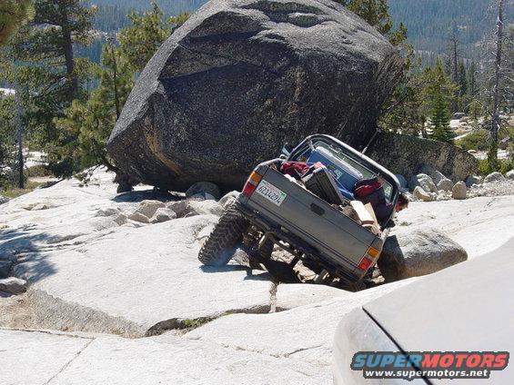 Gray off-road vehicle with a lifted suspension is tilted on rocks, carrying luggage in its bed