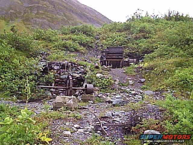 Abandoned mining equipment, including a large wheel and wooden structures, sits in a rocky, overgrown outdoor area with a mountain in the background