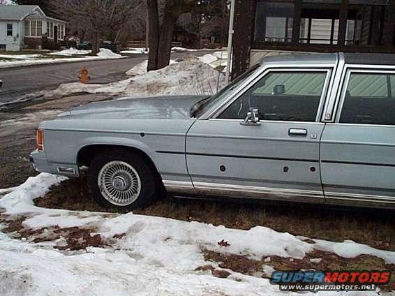 Side view of a light blue Ford sedan parked on a snowy roadside. It has bullet holes along the door