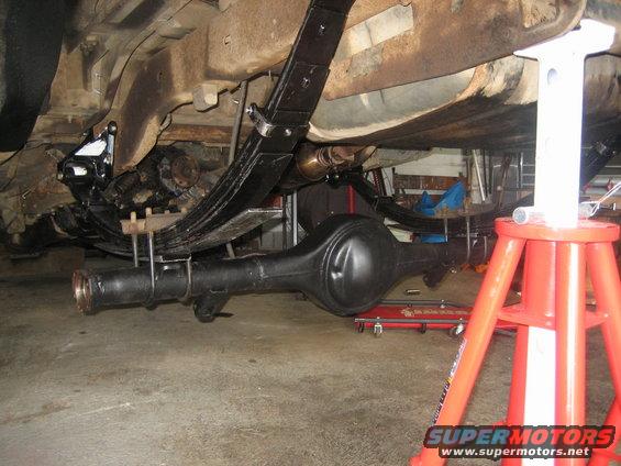 Underside of a vehicle showing the rear axle, leaf springs, and suspension components. The vehicle is supported by jack stands