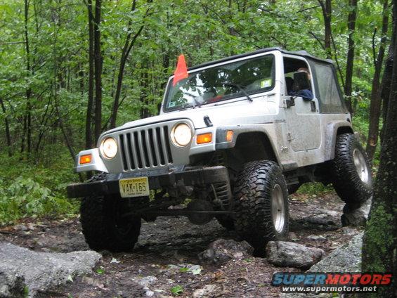 Silver Jeep Wrangler on rocky terrain, front 3/4 view. Orange flag on antenna