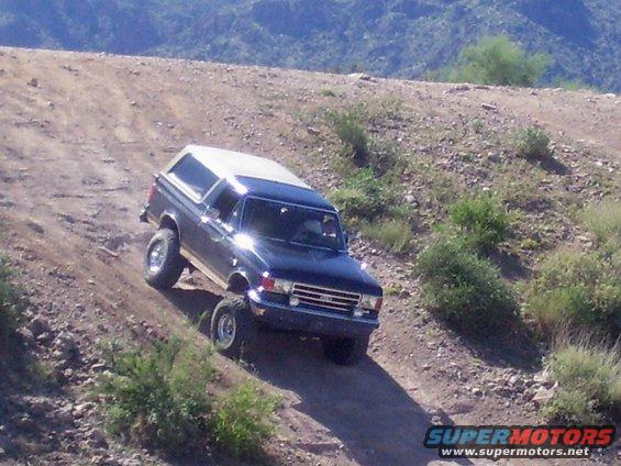 Dark blue Ford Bronco with white top driving up a dirt hill, mountains in background
