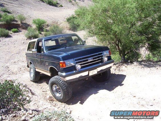 1991 Ford Bronco driving on a dusty, off-road trail with sparse vegetation