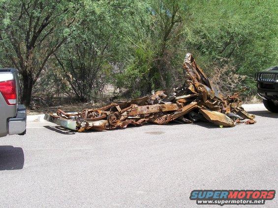 Pile of rusted vehicle frame and parts on the side of the road next to a pickup truck and a black SUV