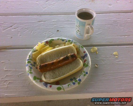 hot dog on a bun with chips and a mug of coffee on a picnic table