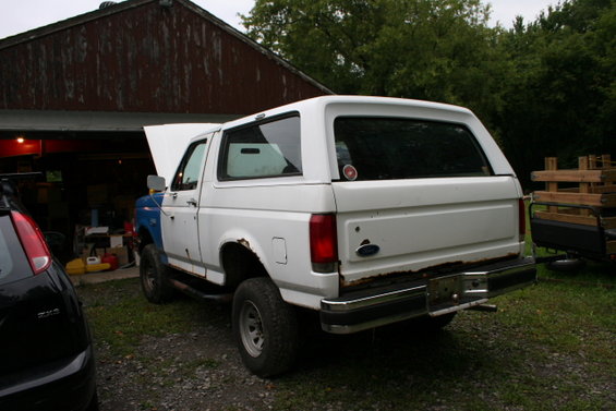 White Ford Bronco with blue front fender, hood up, parked outside a barn
