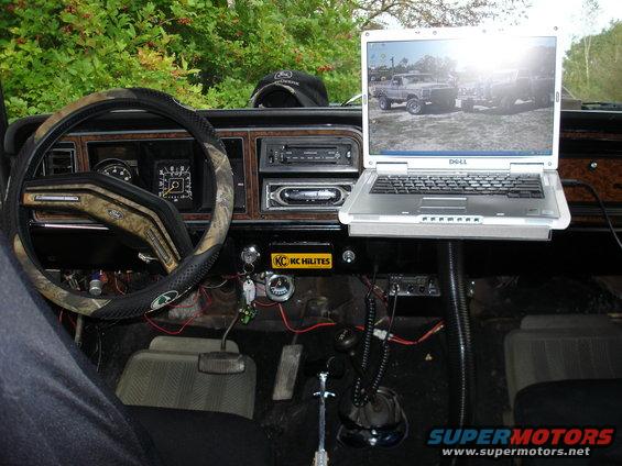 Dashboard of a vehicle with a laptop open on a mount, showing a picture of a truck on its screen. Camouflage steering wheel cover